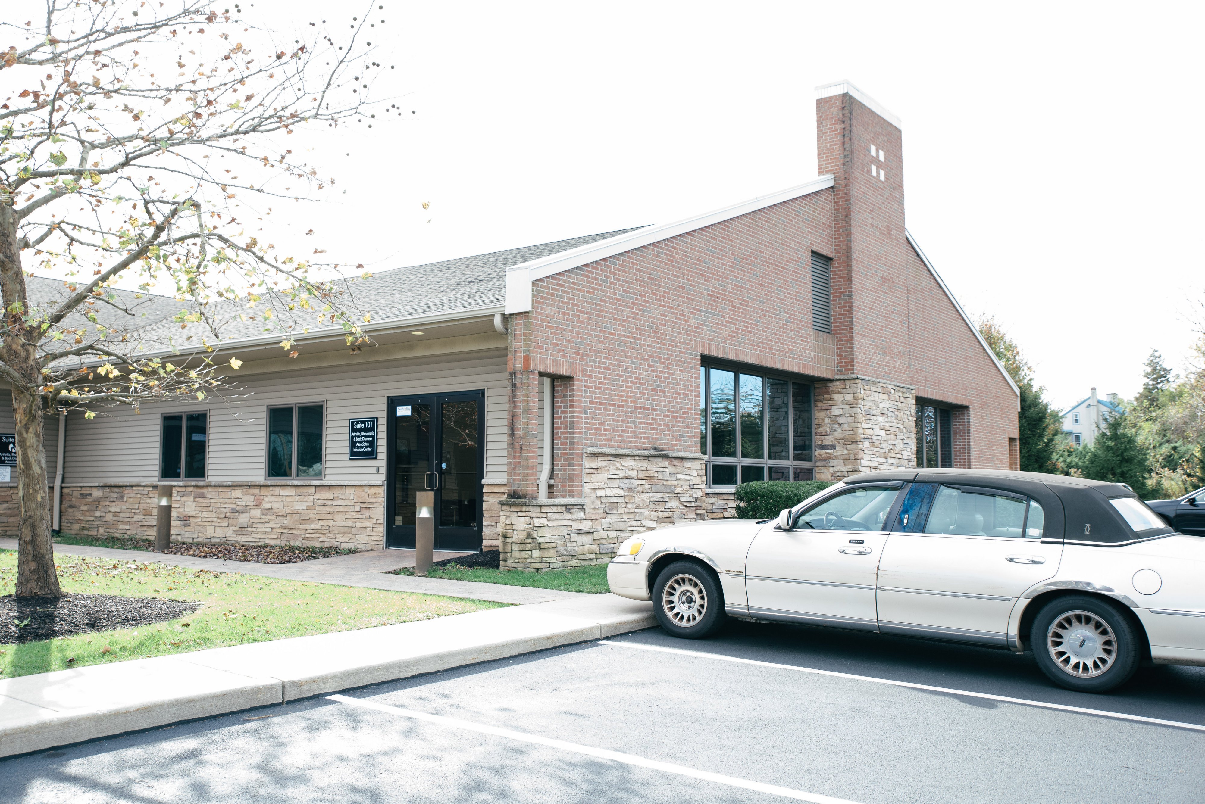 A brick and stone building with large windows, a black sedan parked in front, and a tree in the foreground.